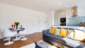 A modern living room with a grey sofa, yellow and grey patterned cushions, a dining table with white chairs, and a kitchen in the background.