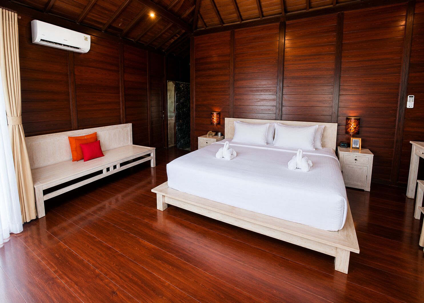 Une chambre d’hôtel avec un plancher en bois, un lit avec une literie blanche, un banc avec des coussins, des lampes de chevet et un appareil de climatisation.