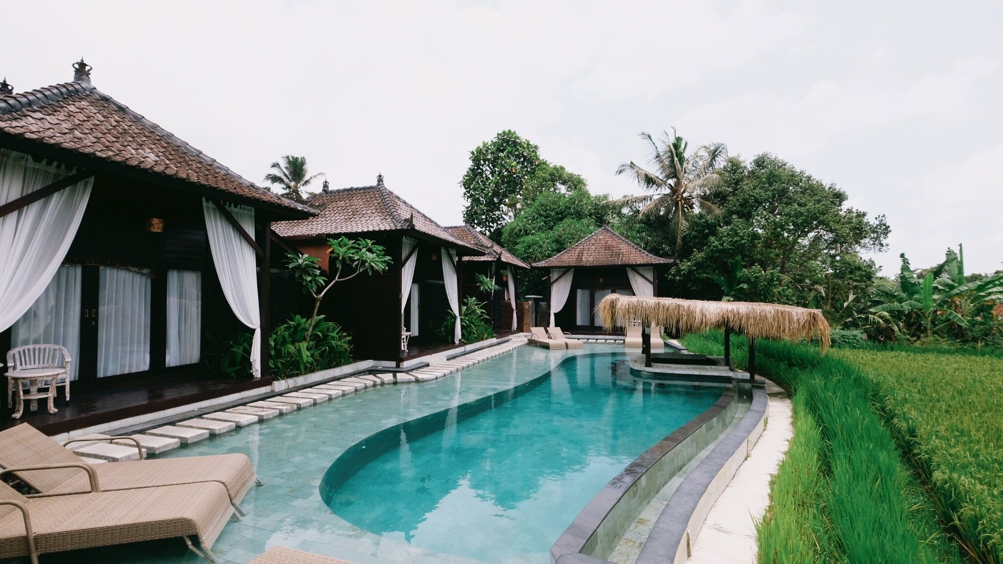A swimming pool with lounge chairs and thatched-roof huts, surrounded by lush greenery.