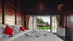 Een groot bed met witte lakens en rode kussens, een houten plafond en uitzicht op een tuin door een open deur.
