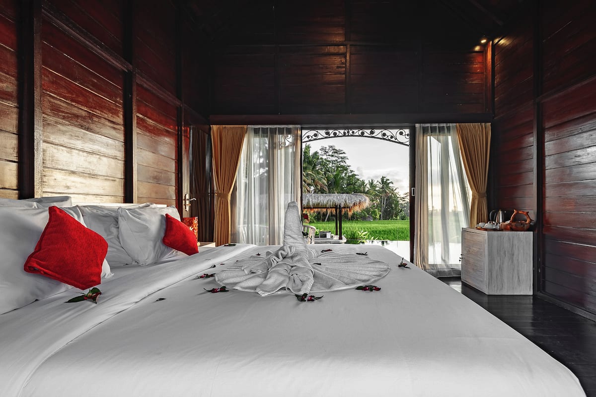 Un grand lit avec du linge de lit blanc et des oreillers rouges, un plafond en bois et une vue sur un jardin par une porte ouverte.