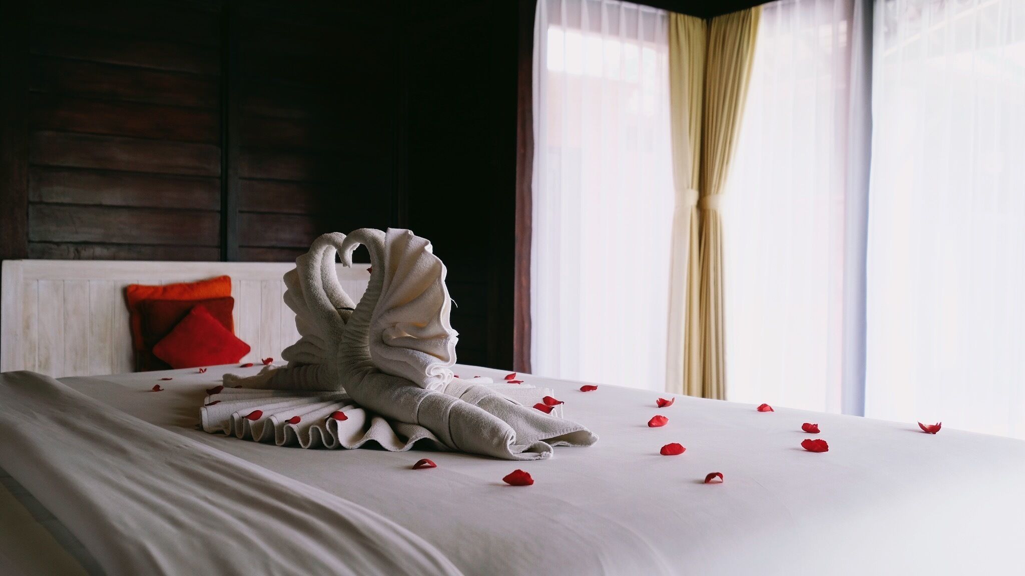 A neatly made bed with a swan-shaped towel arrangement and scattered rose petals.