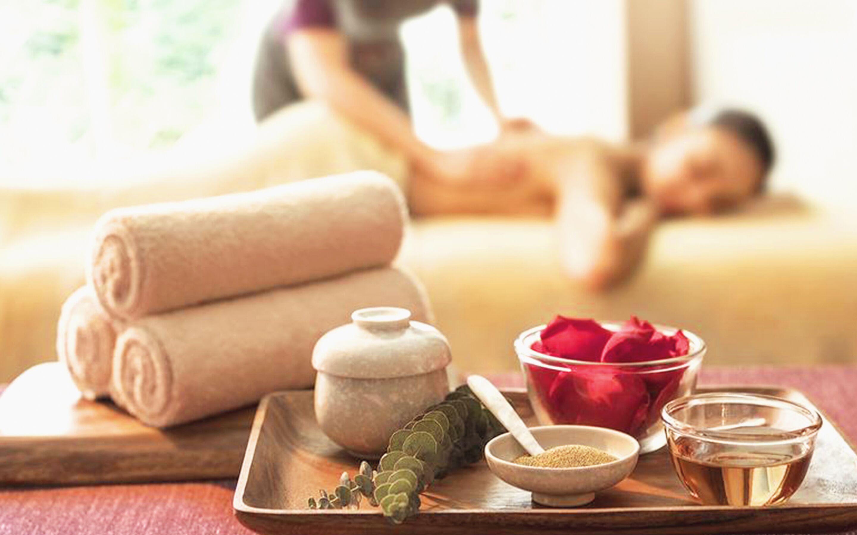 Un ambiente de spa con una camilla de masaje, toallas enrolladas, un cuenco de pétalos de rosa, un cuenco de hierbas y una bandeja con un recipiente de aceite y un frasco pequeño.
