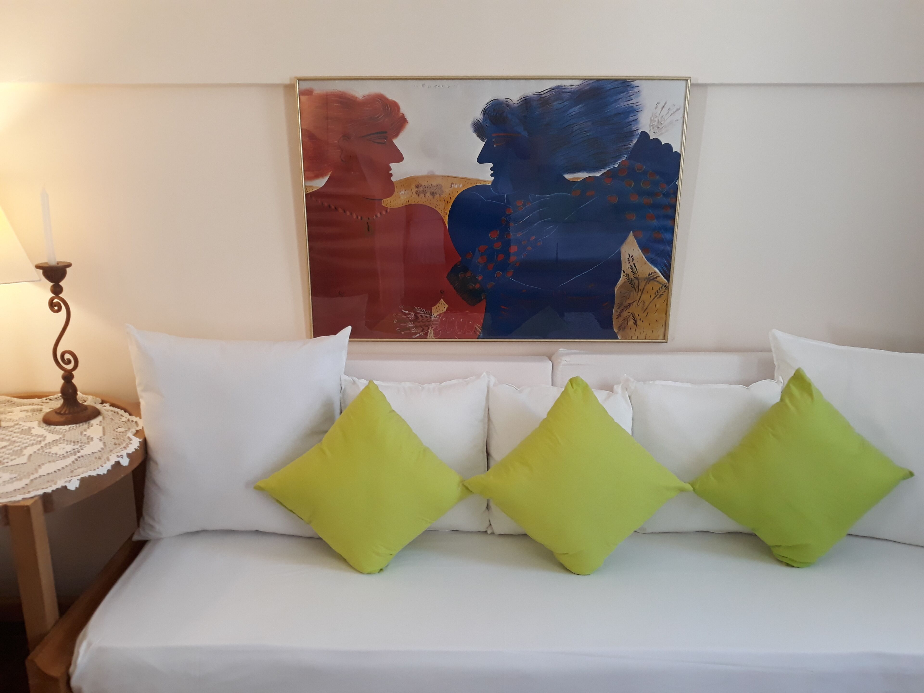 Uma cama com almofadas brancas e verdes, um quadro abstrato emoldurado acima e uma pequena mesa com uma luminária à esquerda.