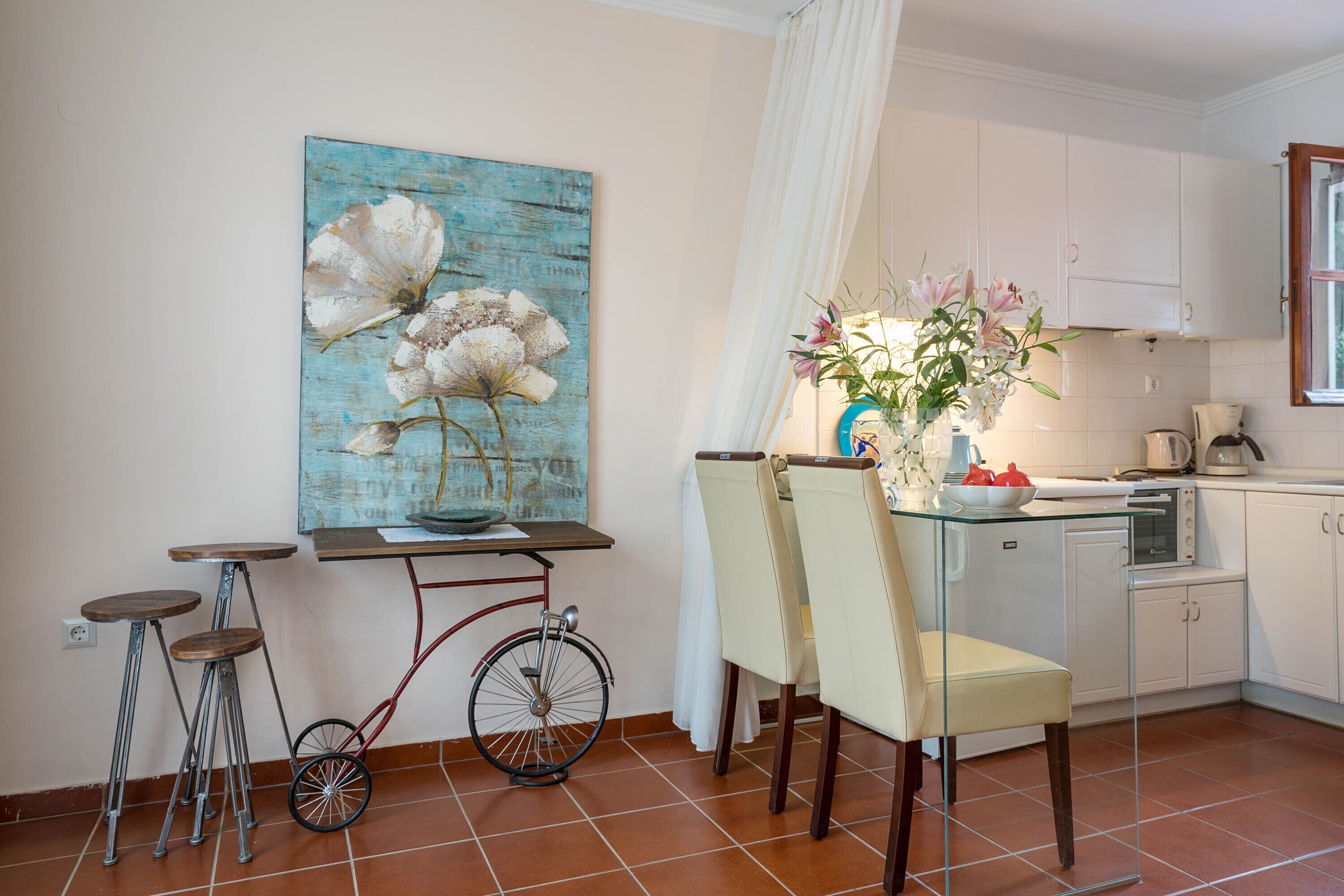 Eine Küche mit weißen Schränken, einem Esstisch mit Stühlen, einem Fahrrad und einem Blumengemälde an der Wand.