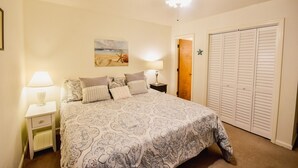 Une chambre à coucher comprenant un lit, des tables de chevet, un ventilateur de plafond, un tableau sur le thème de la plage et un placard.