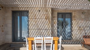 Een buitenpatio met een houten tafel, witte stoelen, een stenen muur en een pergola erboven.
