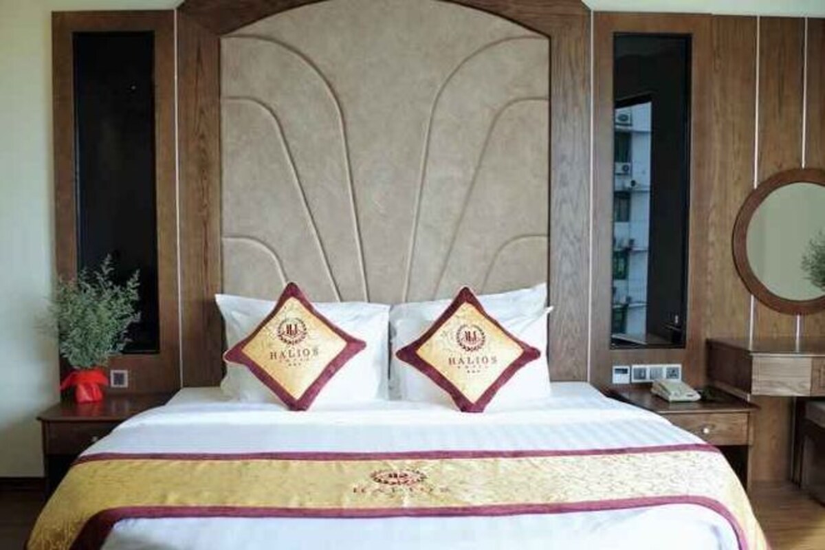 A hotel room with a large bed, two pillows with a logo, a wooden headboard, a bedside table with a phone, and a mirror.