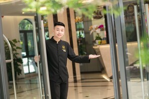 A person in a black uniform standing in a modern lobby with glass doors, a reception desk, and indoor plants. - Halios Hotel Halong (Ha Long)