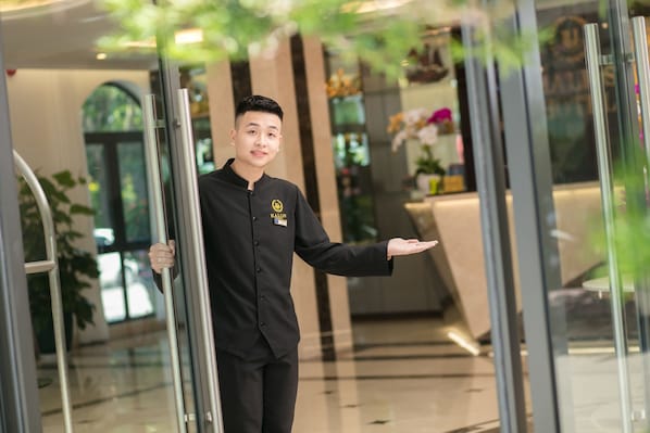 Une personne vêtue d’un uniforme noir se tient dans un hall d’entrée moderne, doté de portes vitrées, d’un comptoir d’accueil et de plantes d’intérieur.
