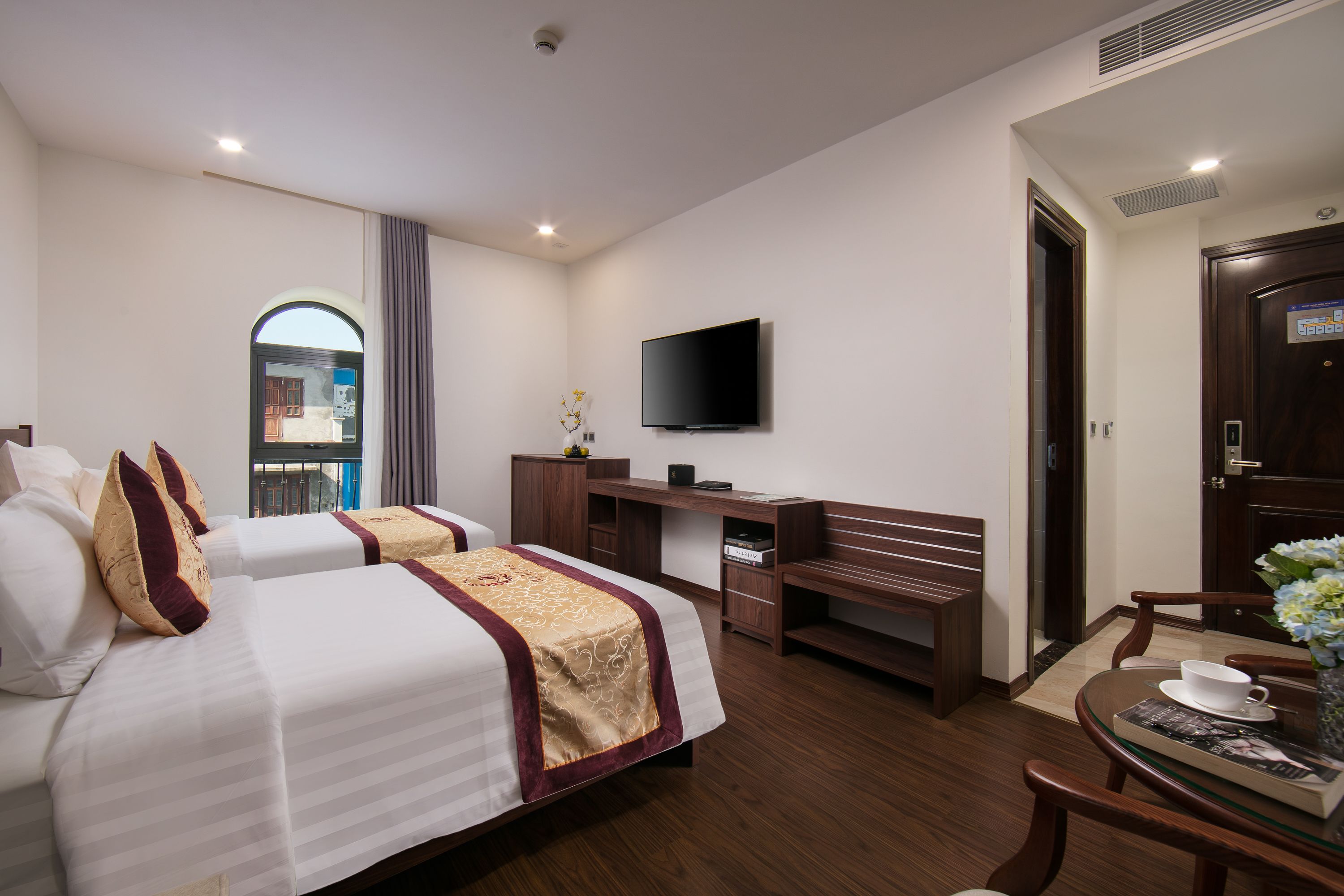 A hotel room with two beds, a desk, a chair, a television, and a small table with a cup and a book.