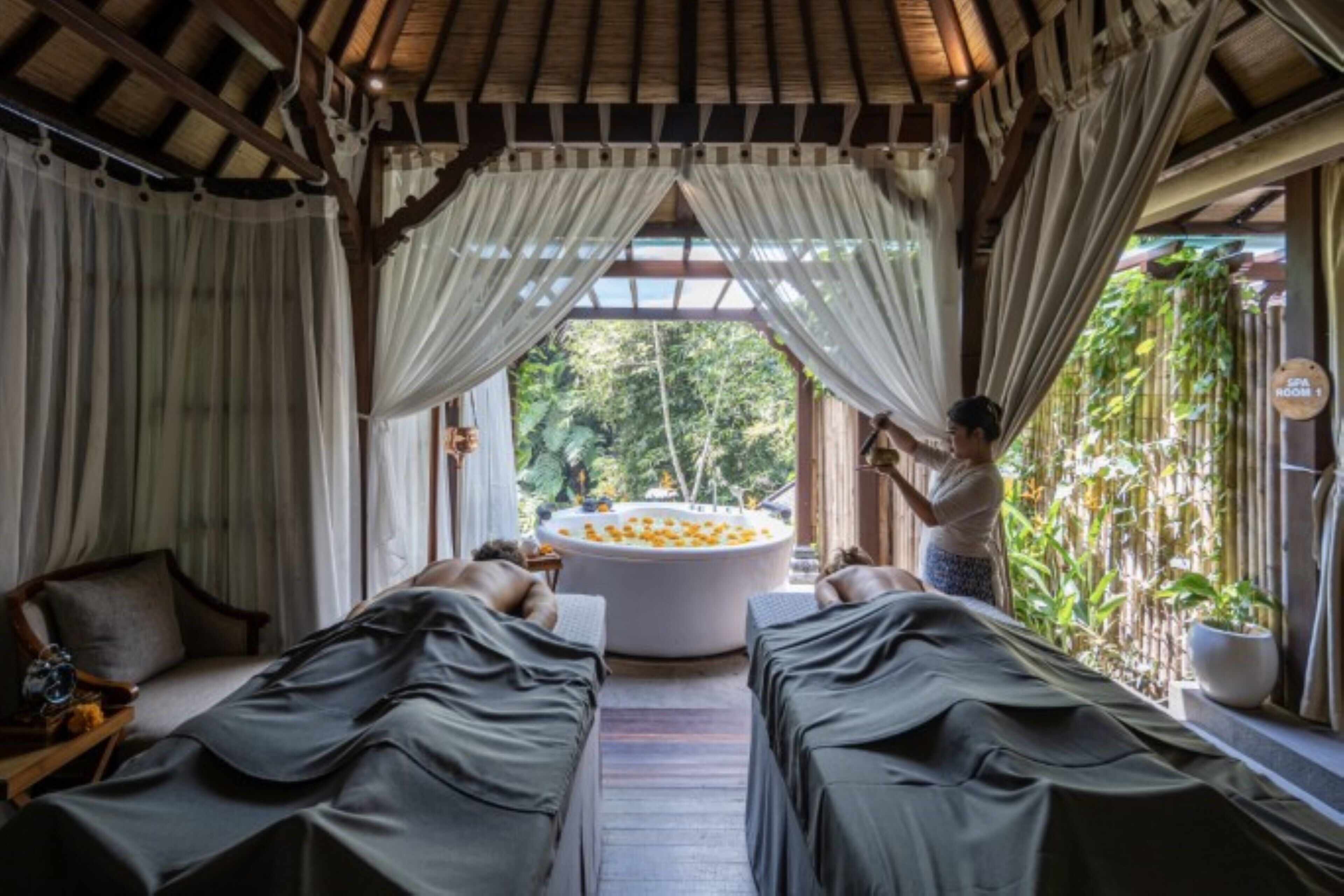 A spa room with two massage tables, a hot tub with orange flowers, and a view of greenery outside.