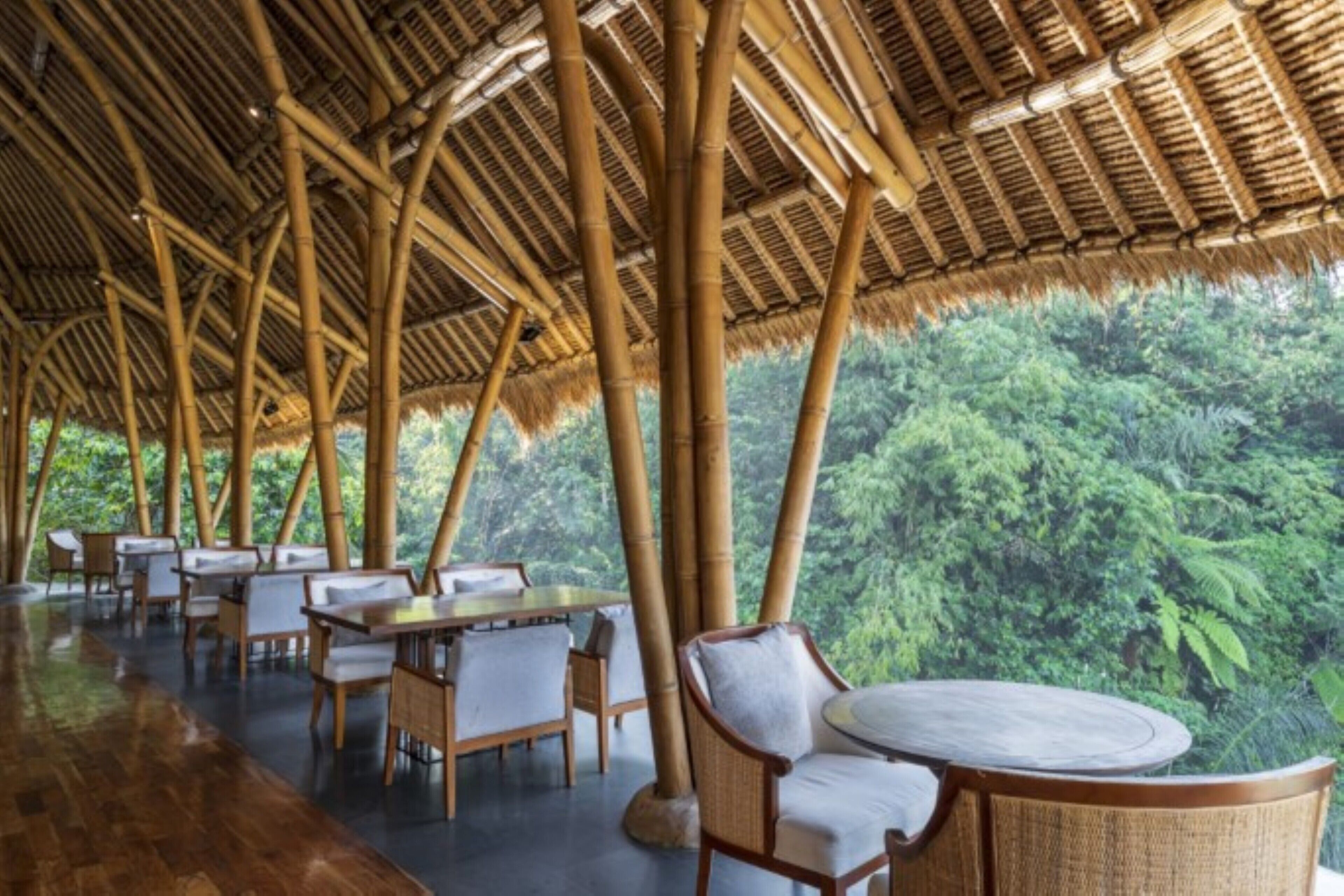 An open-air restaurant with bamboo ceilings and tables, surrounded by lush greenery.