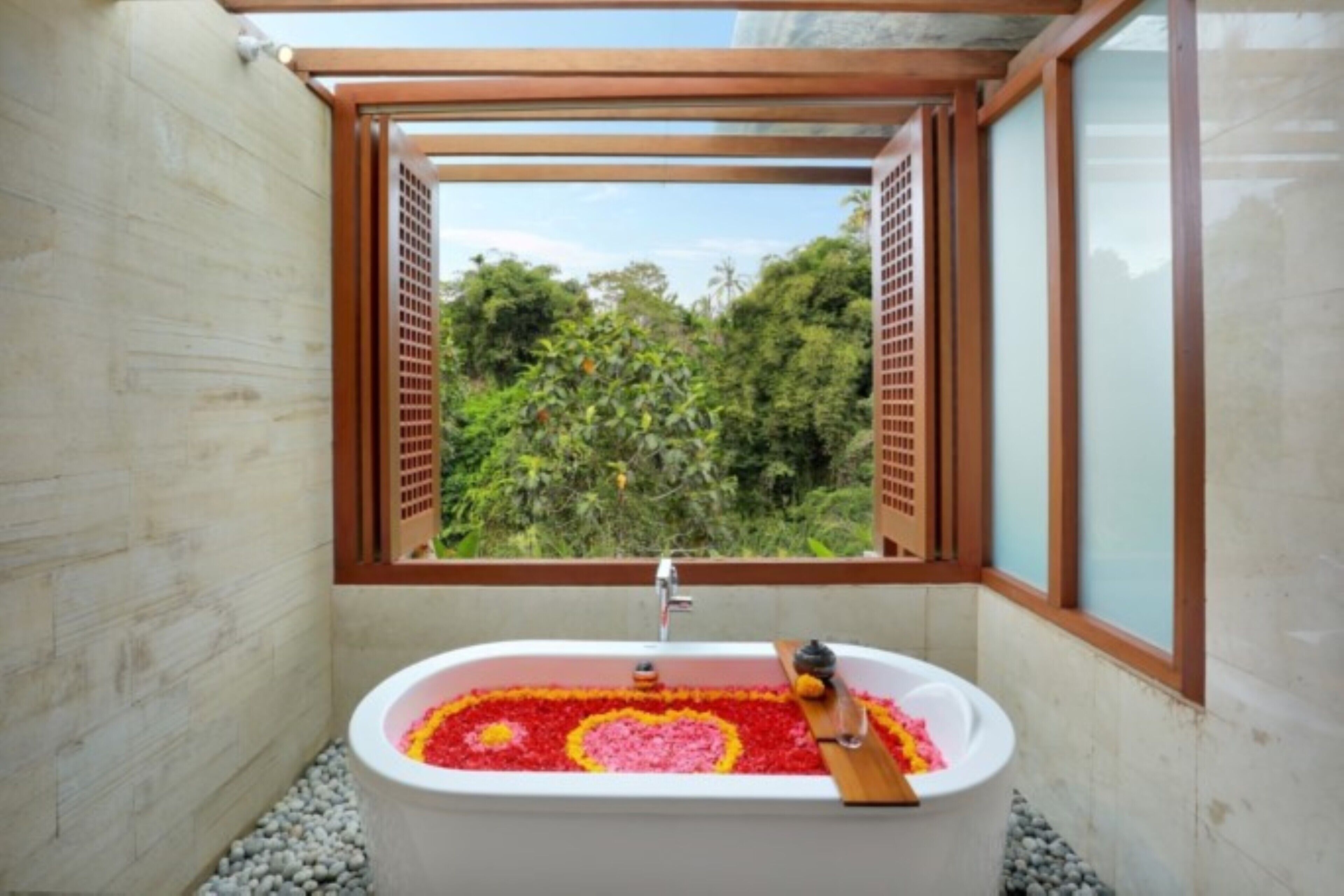 A bathroom with a bathtub filled with rose petals, a window with wooden shutters, and a view of greenery outside.