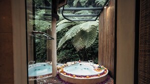 A glass-enclosed spa area with two hot tubs surrounded by flower petals, set against a backdrop of lush greenery.