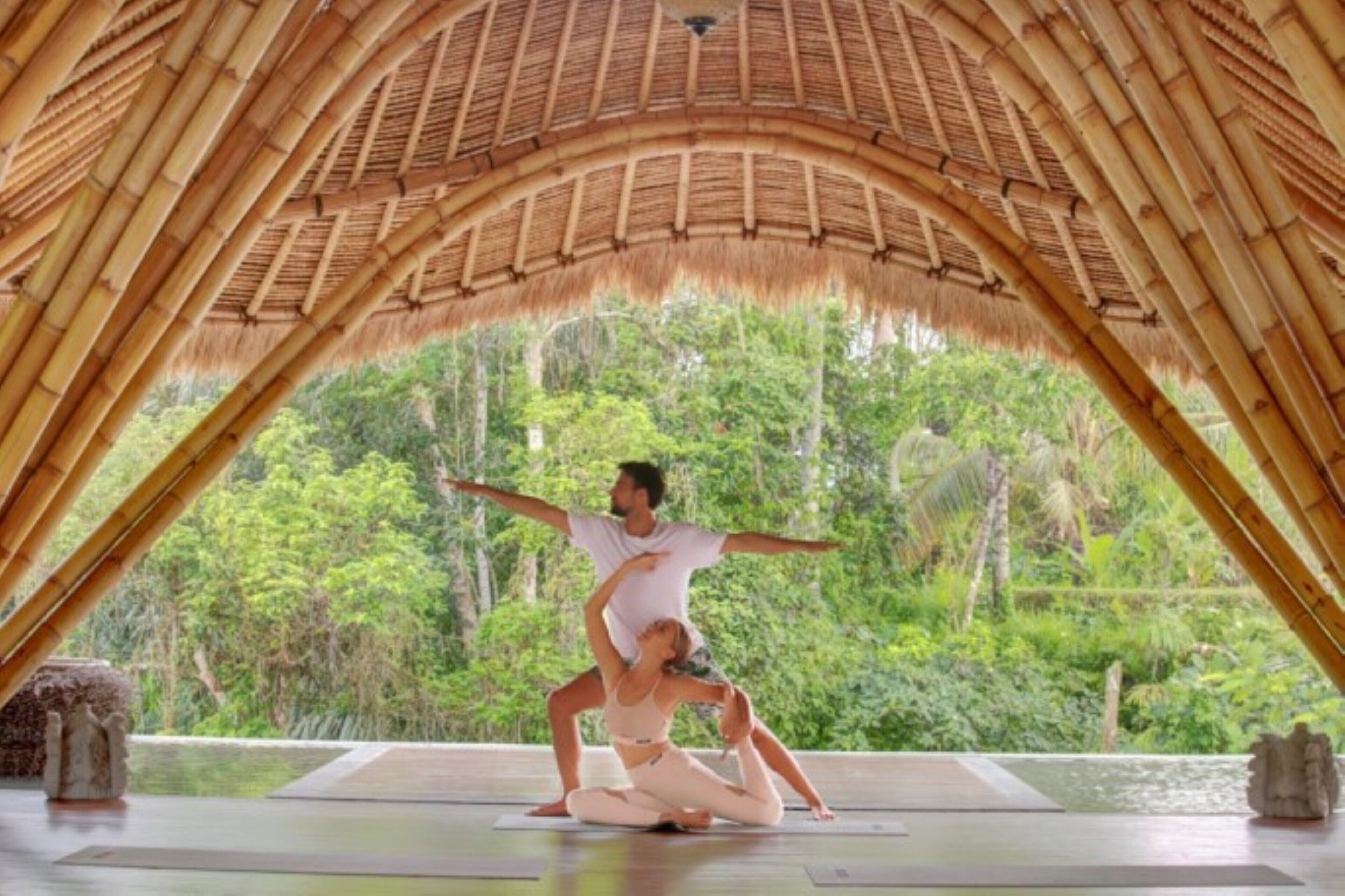Una coppia che pratica yoga in una stanza con soffitto in bambù e vista su una vegetazione rigogliosa.