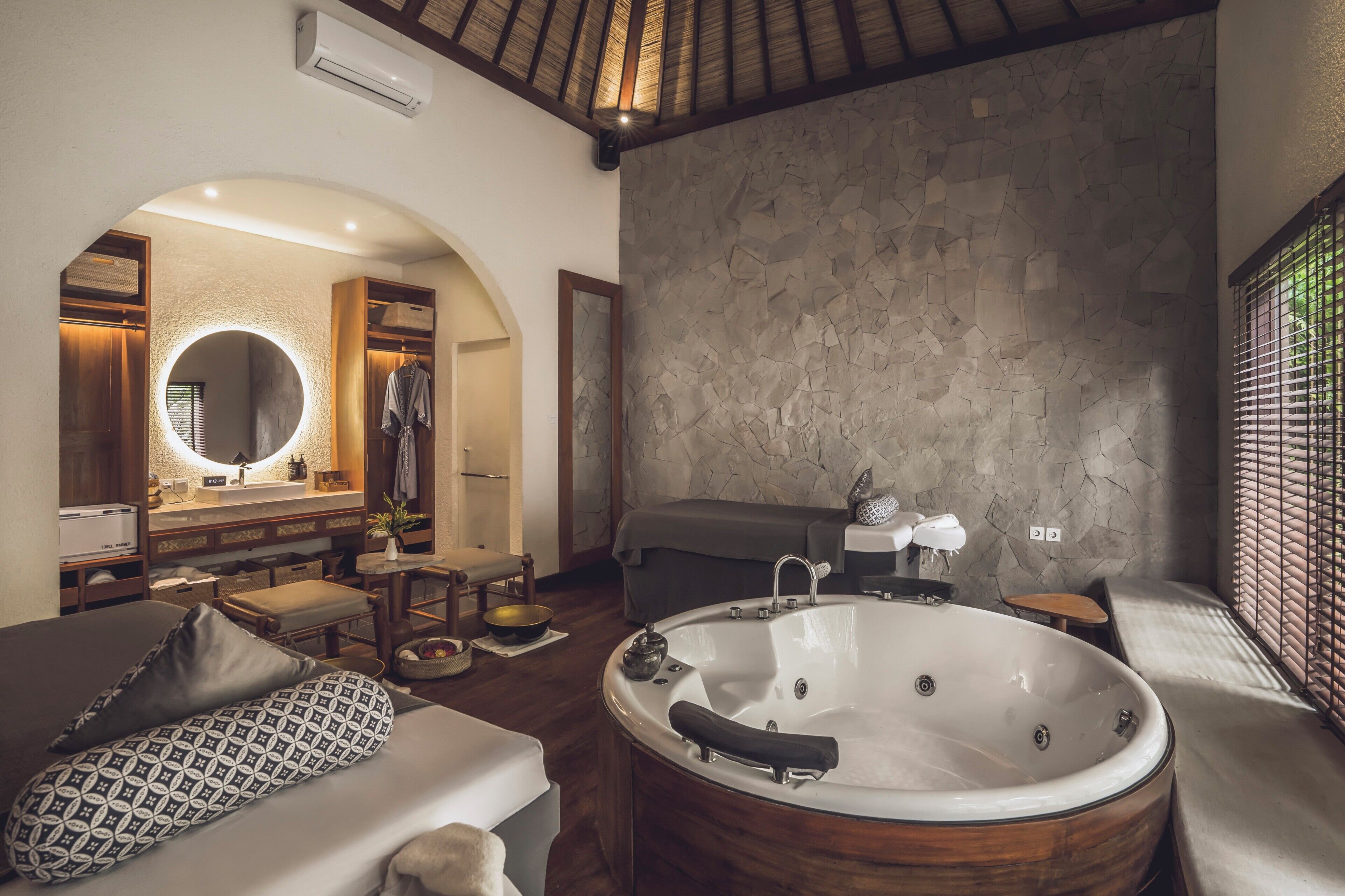 Ein geräumiges Badezimmer mit einer großen Badewanne, einem runden Spiegel, einer Steinwand und einer Holzbalkendecke.