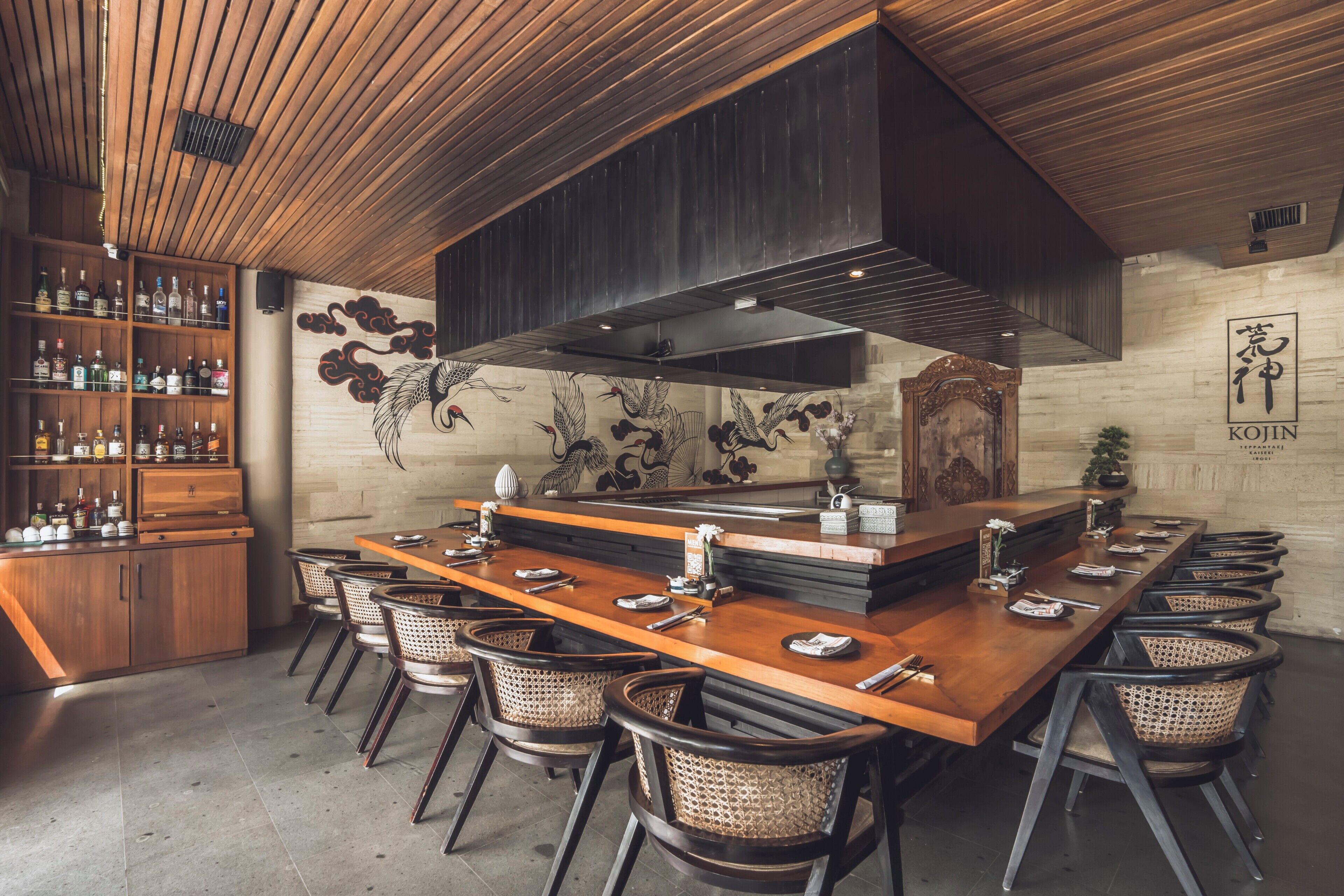 A restaurant with a long wooden bar, wicker chairs, and a modern kitchen.