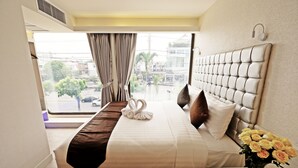 A hotel room with a large bed, a headboard, a bedside table with a vase of flowers, and a view of the city through the window. - Justice Hotel (Bangkok)