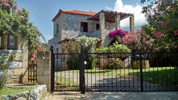 A stone house with a red-tiled roof, a gated entrance, and a well-maintained garden with flowering plants. - First floor apartment with beautiful mountain and sea view near the sea (Στούπα)