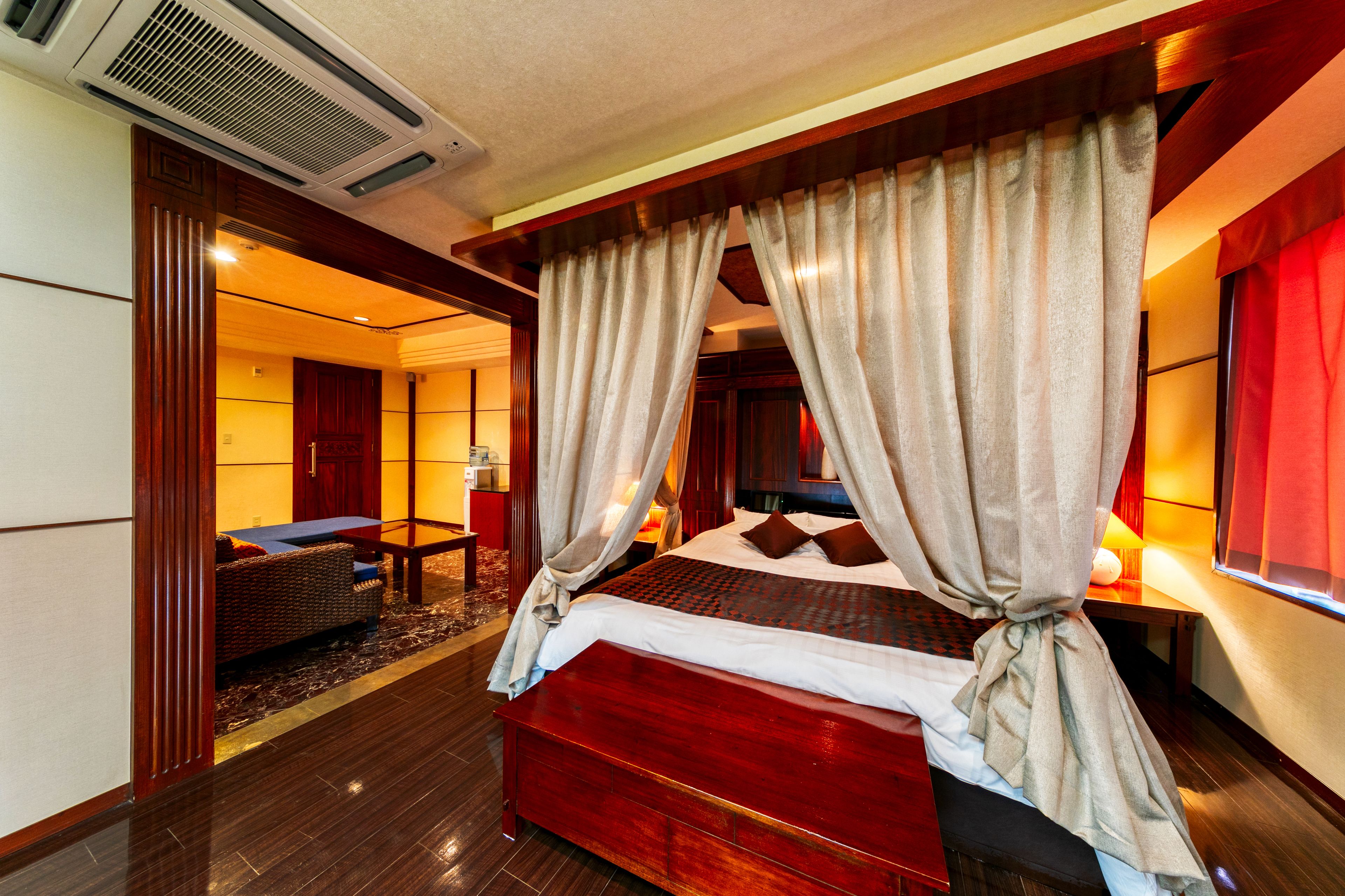 A spacious bedroom with a large bed, a sitting area with a sofa and table, and a separate room with a desk and chair.