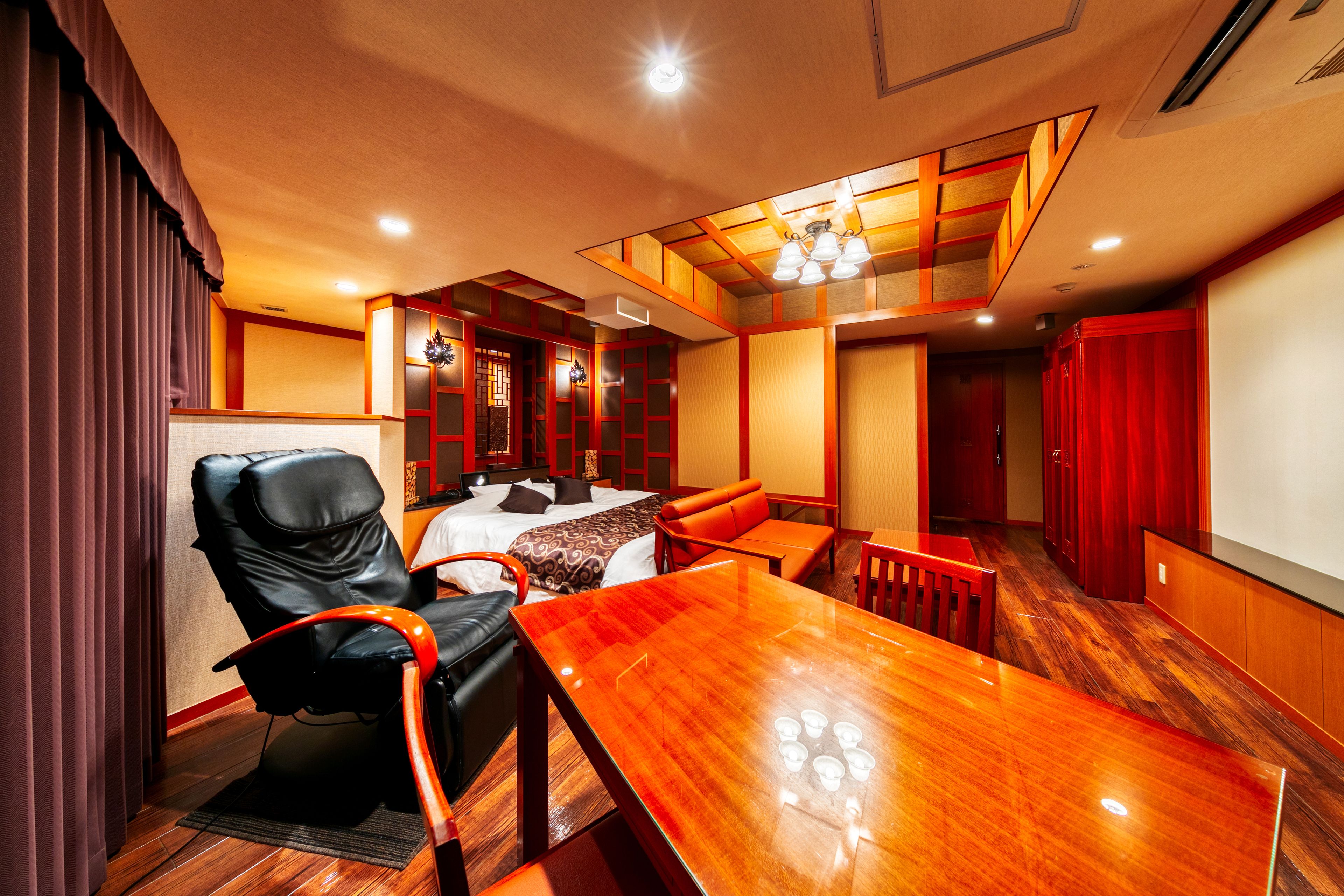 A hotel room with a wooden dining table, a black leather chair, a sofa, and a bed with a patterned bedspread.