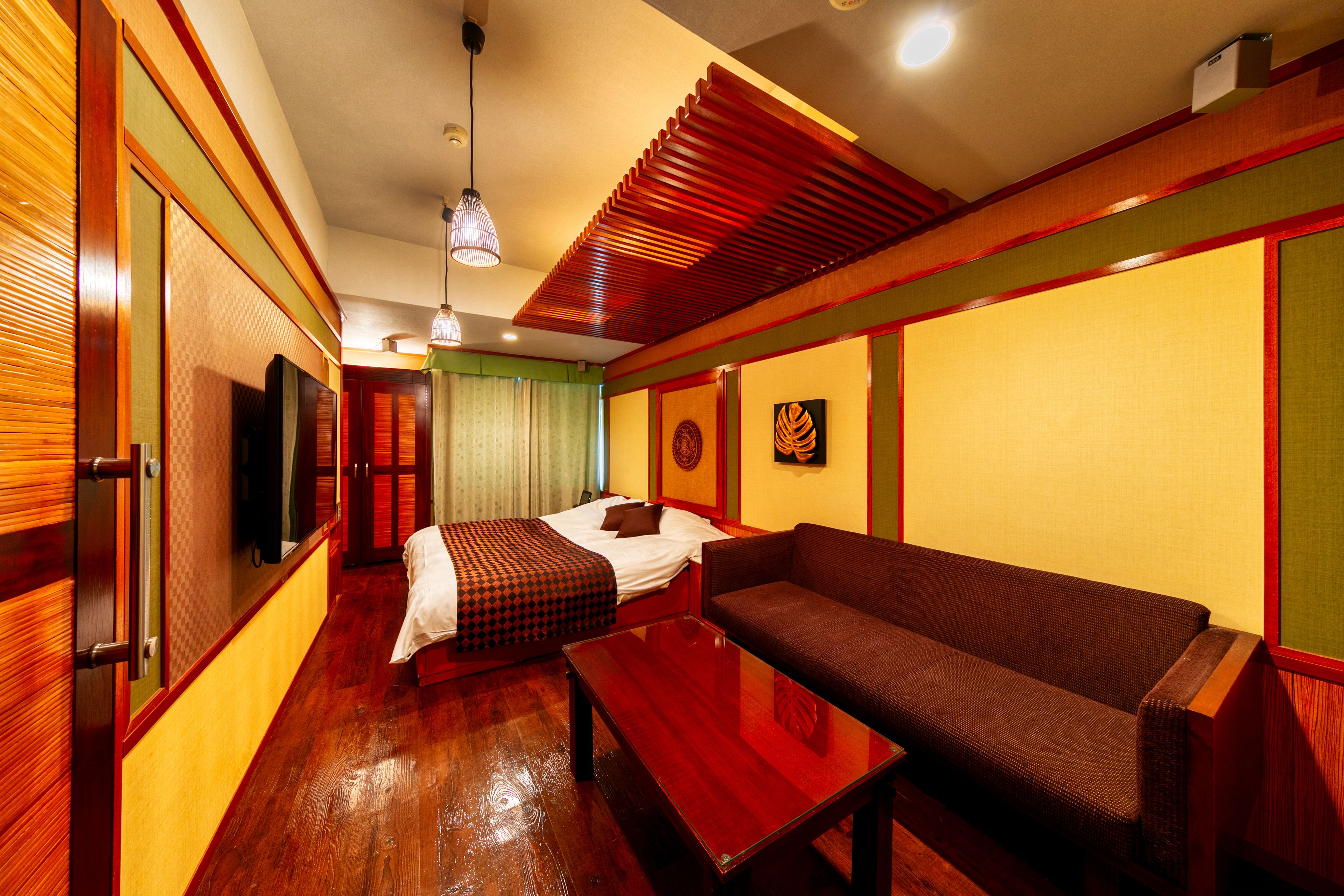 A hotel room with a wooden floor, a bed with a patterned bedspread, a sofa, a wooden table, and a television.
