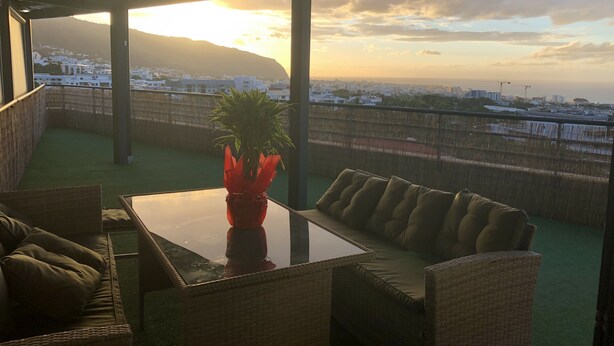 Ein Balkon mit Korbmöbeln, einem Glastisch und einem roten Blumenvasen mit Pflanze.