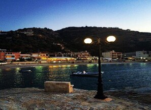 A coastal town at dusk with a stone bench, a lamp post, and boats on the water. - Dream apartment in a top location directly on the beach, with beautiful sea views (Λυγαριά)