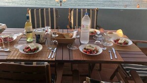 A table set for a meal with plates of food, glasses, and bottles, overlooking a coastal area with people in the water and a rocky outcrop in the distance. - Dream apartment in a top location directly on the beach, with beautiful sea views (Λυγαριά)