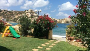 A coastal garden with a children's play area, including a slide, and a view of the sea and buildings in the background. - Dream apartment in a top location directly on the beach, with beautiful sea views (Λυγαριά)