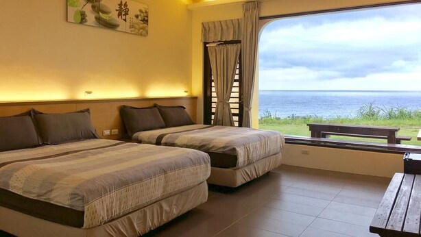 A hotel room with two beds, a large window with a view of the sea, a wooden bench, and a wall-mounted artwork. - White Sand Beach 32 (Lyudao)
