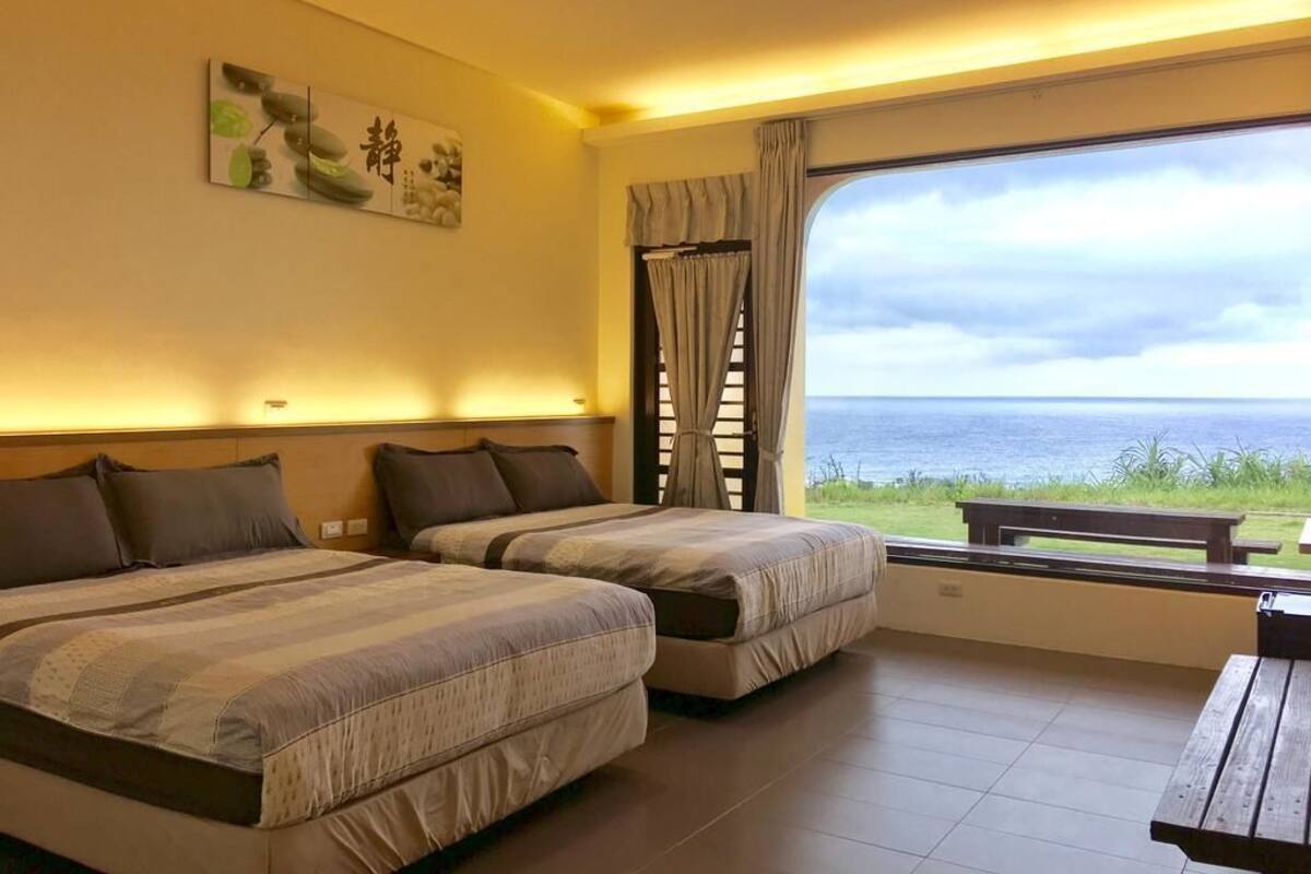 Habitación de hotel con dos camas, un ventanal con vistas al mar, un banco de madera y una obra de arte colgada en la pared.