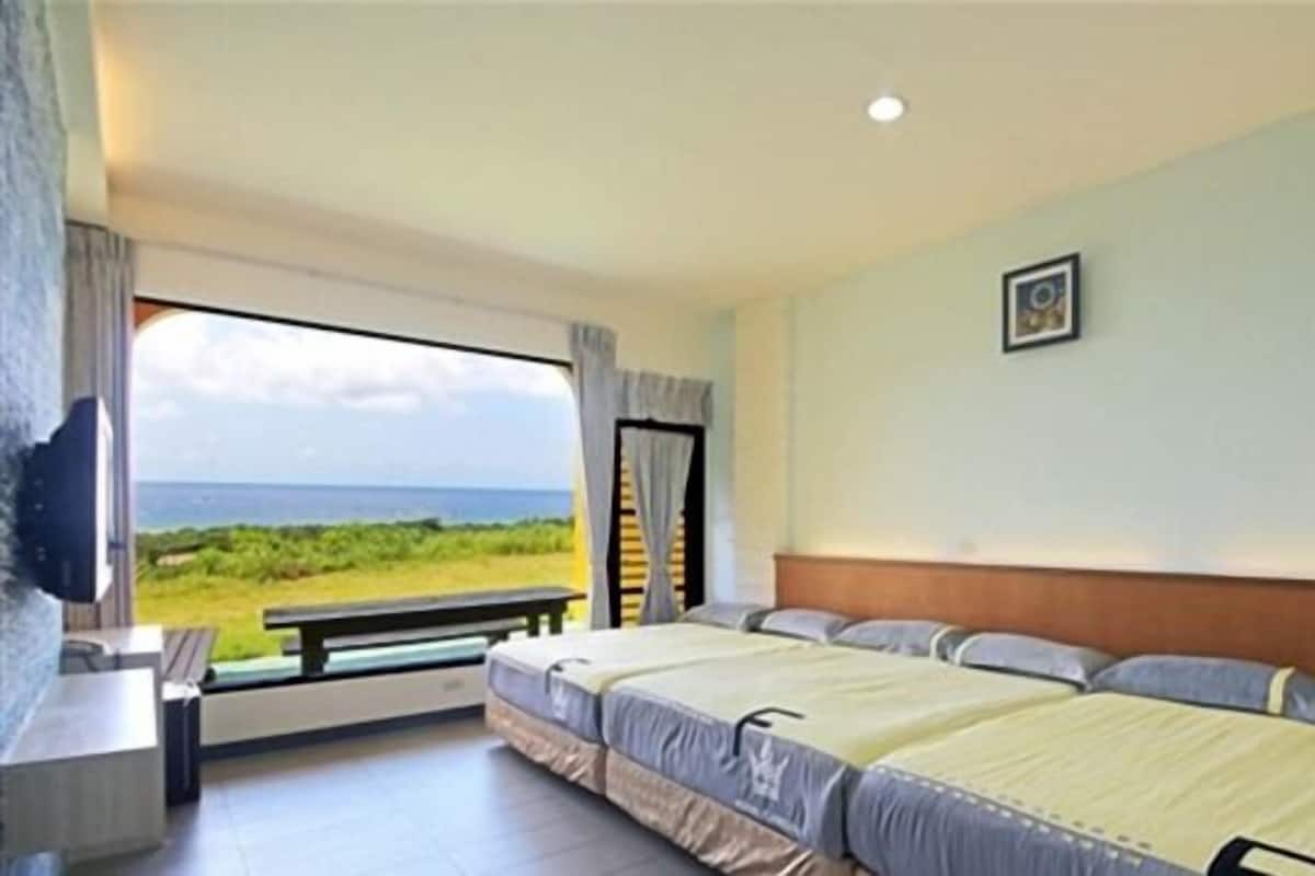 Habitación de hotel con dos camas, un escritorio y un amplio ventanal que ofrece vistas a la playa y al océano.