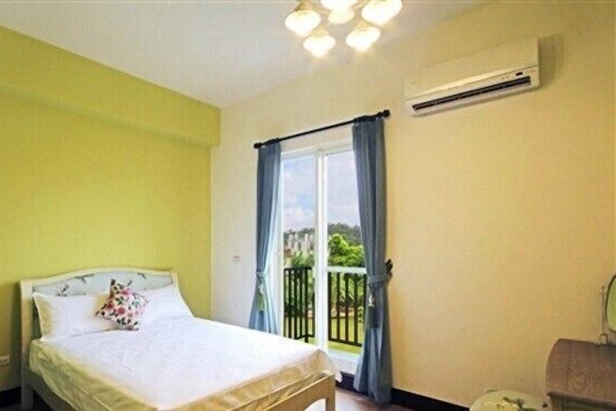A bedroom with a bed, a window with curtains, an air conditioning unit, and a view of the outside.