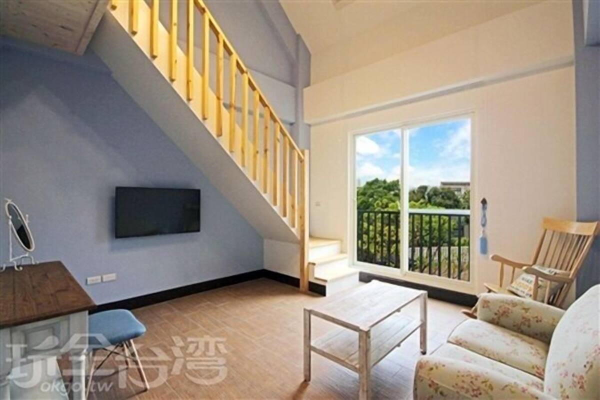 A spacious living area with a wooden staircase, a flat-screen TV mounted on the wall, a wooden coffee table, and a floral sofa.
