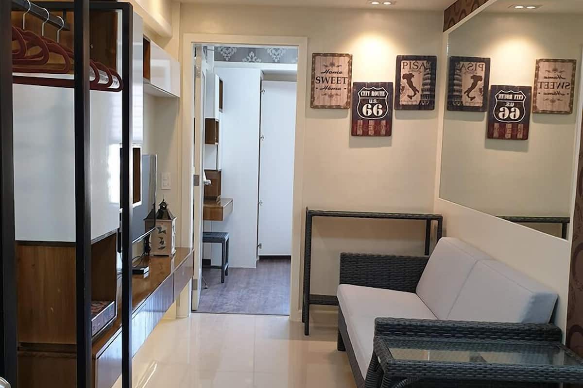 A modern living room with a sofa, a glass coffee table, and a ceiling fan. There are framed signs on the wall and a door leading to another room.