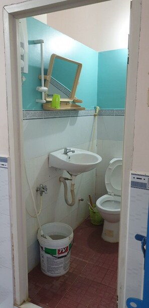 A small bathroom with a sink, toilet, and a mirror. There is a bucket on the floor, and the walls are tiled. - Nha Tro Kim Anh (Long Xuyen)