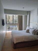 Quarto com uma janela grande, cama com roupa de cama branca e cabeceira moderna.