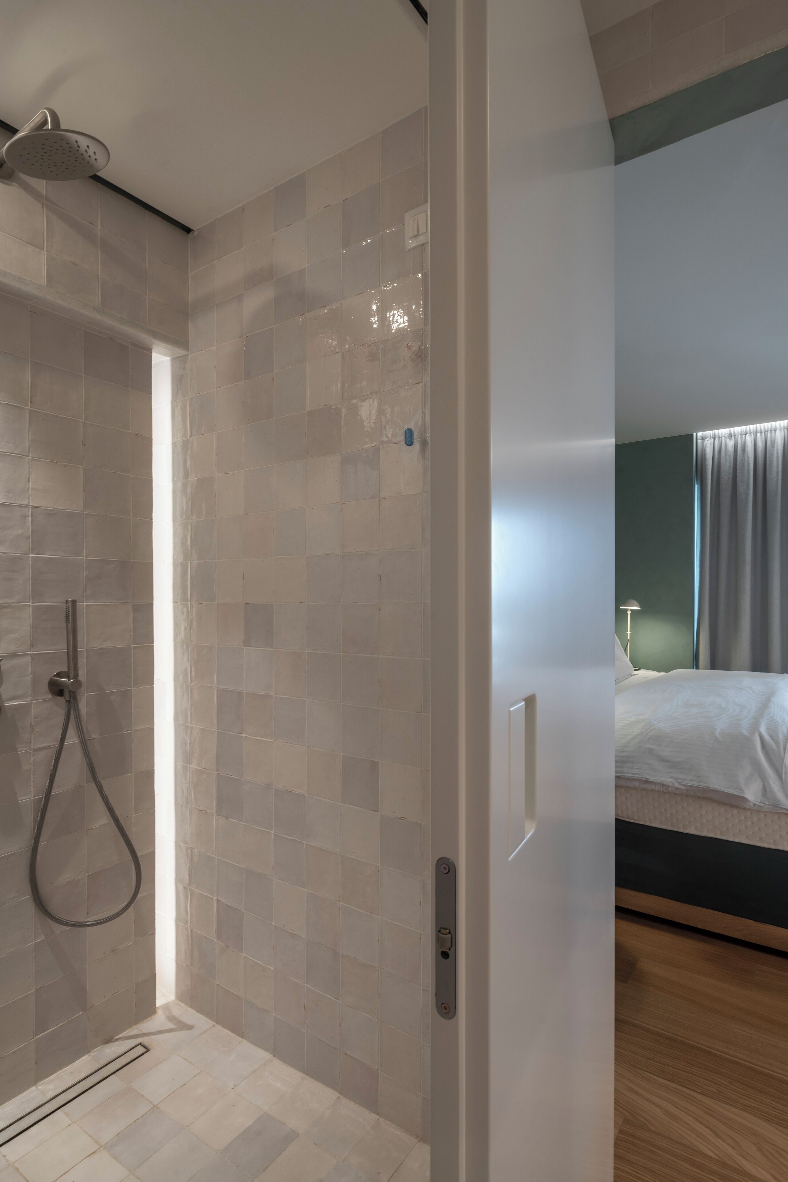 Une salle de bain moderne avec une douche, des murs carrelés et une porte visible donnant sur une chambre à coucher équipée d’un lit et de rideaux.