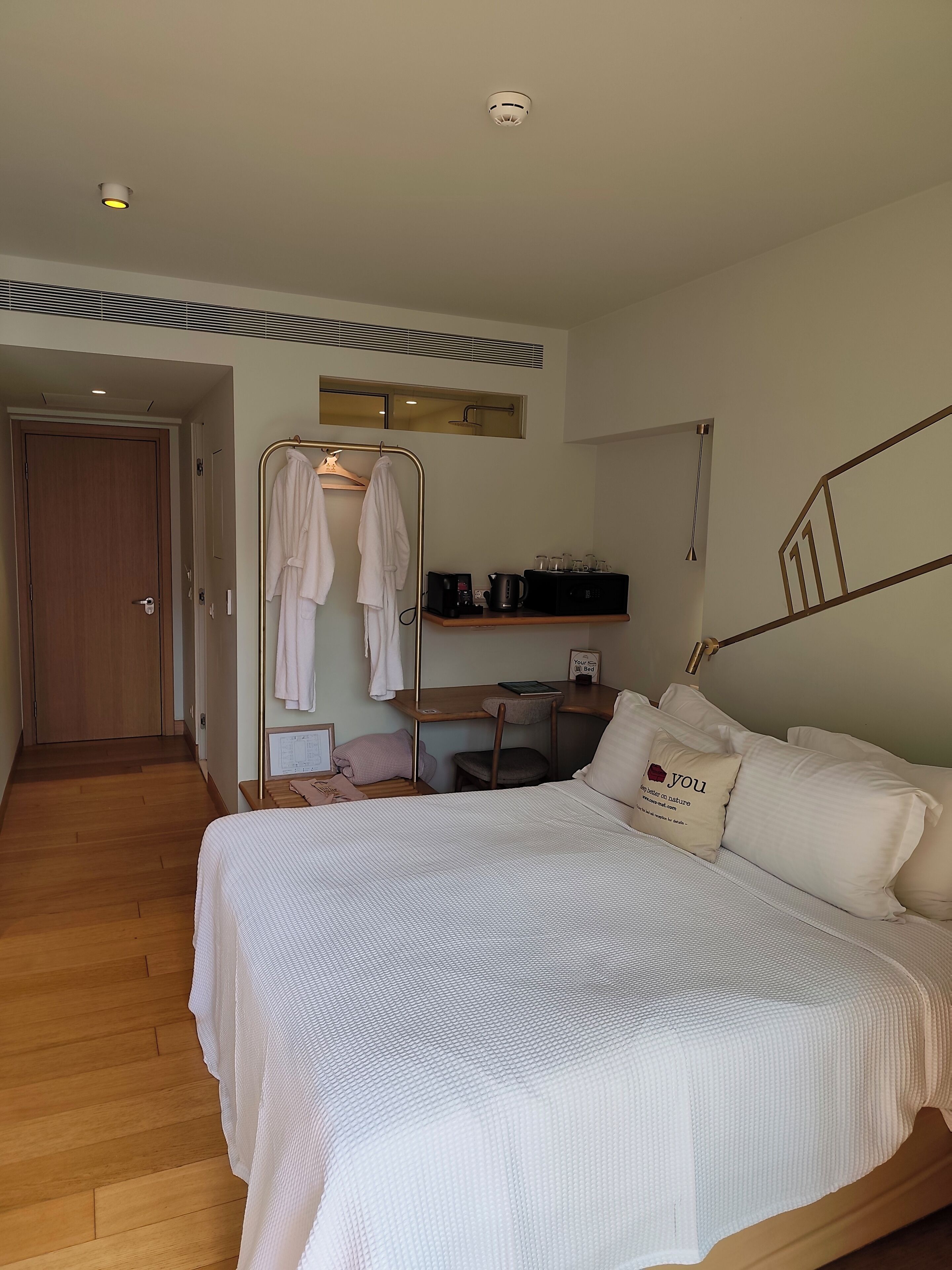 A bedroom with a large bed, a wooden desk, a chair, a hanging robe rack, and a small table with a kettle and cups.