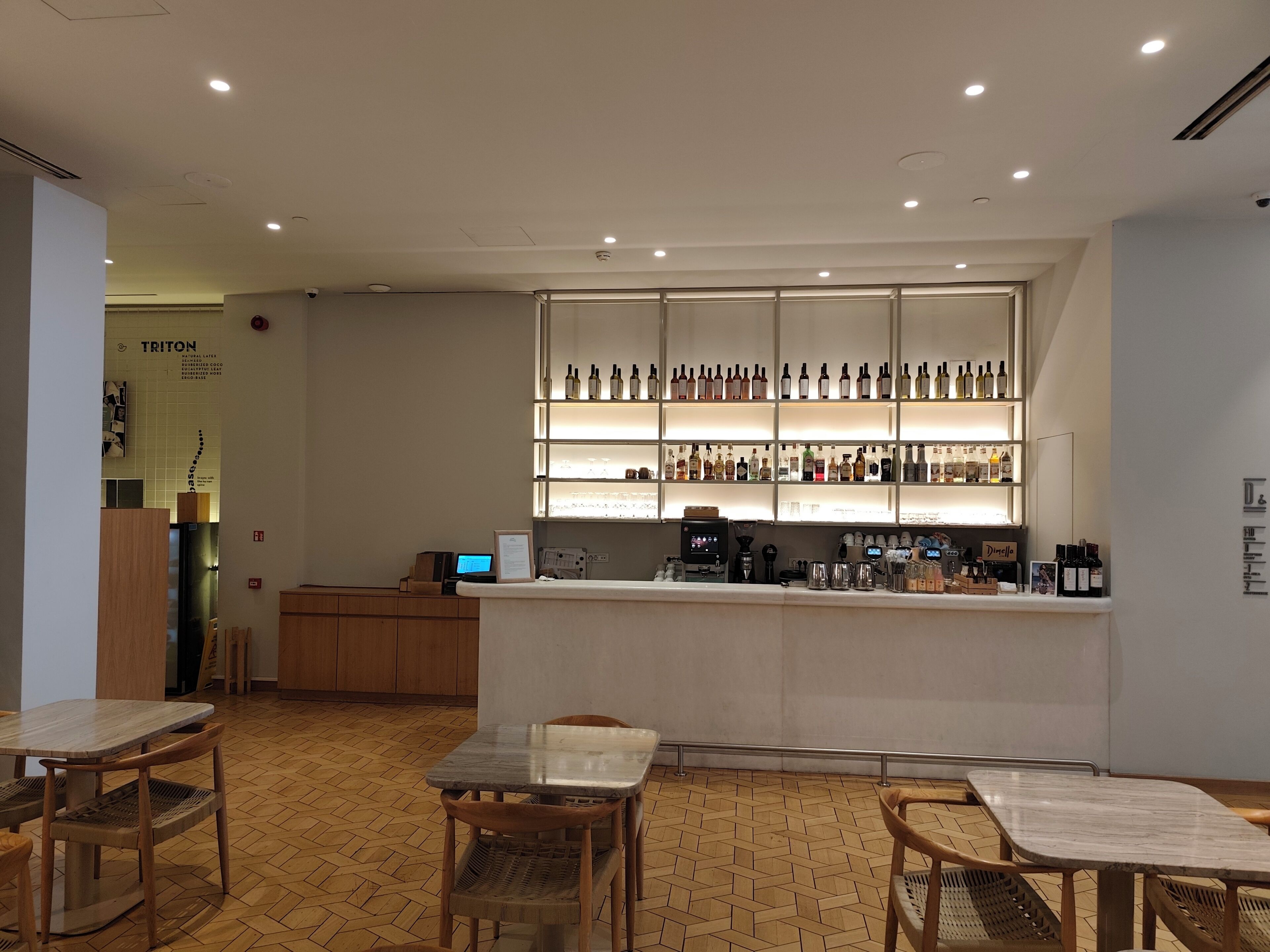Zona de bar con mostrador, estanterías llenas de botellas y una máquina de café. Hay mesas y sillas de madera.