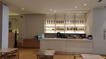 Zona de bar con mostrador, estanterías llenas de botellas y una máquina de café. Hay mesas y sillas de madera.