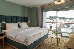 Ett modernt hotellrum med en stor säng, utsikt över staden, ett litet bord med en vas med blommor och en balkong med en stol.