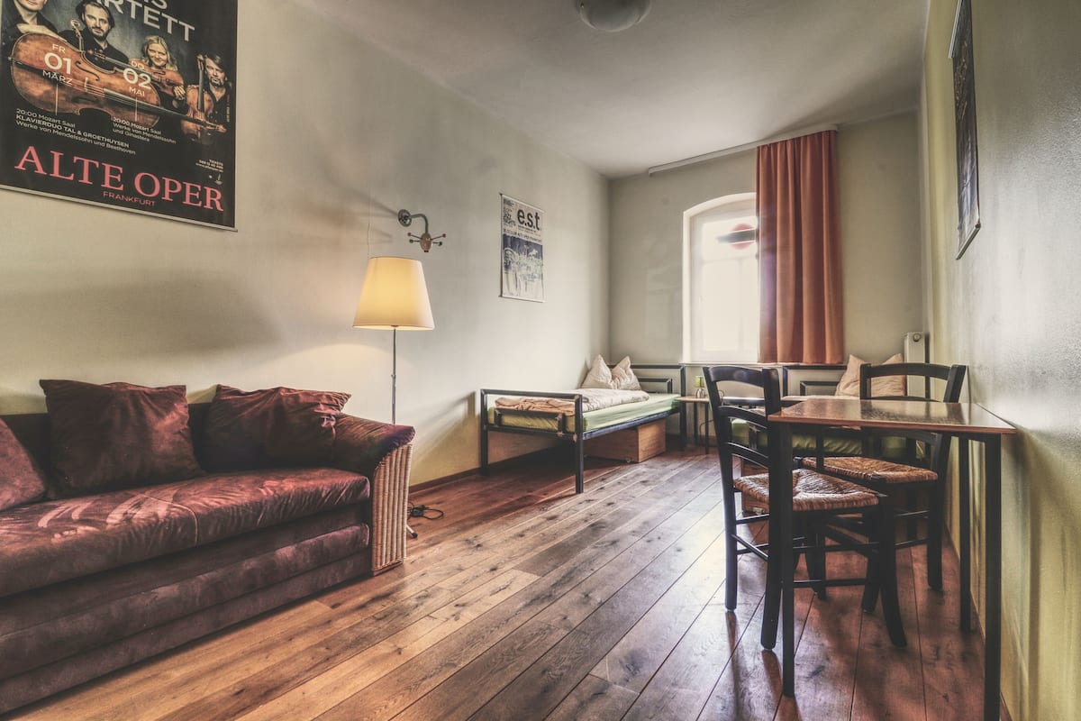 A cozy room with a wooden floor, a sofa, a small table with chairs, a lamp, and a poster on the wall.