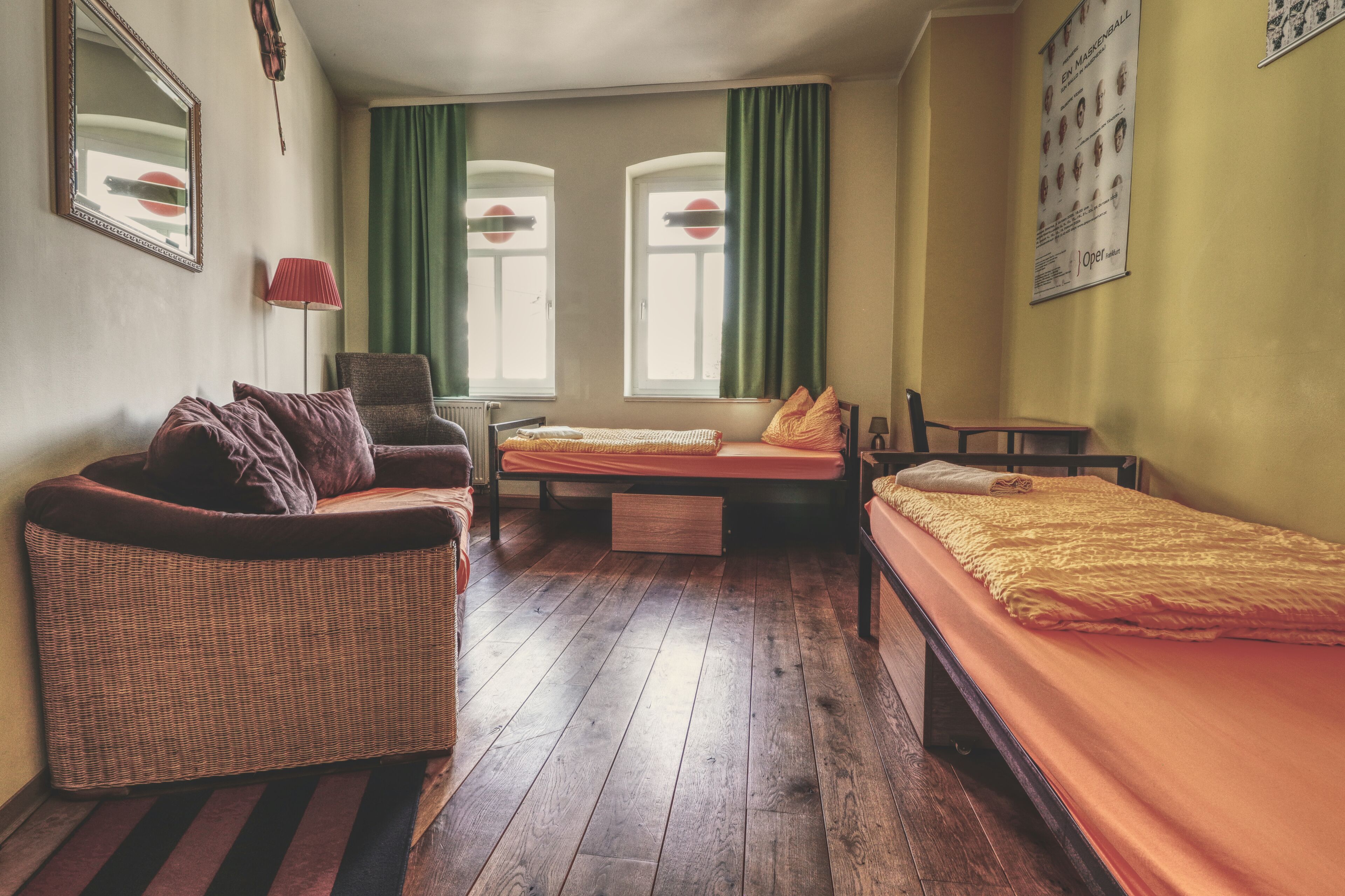 Una habitación con un sofá de mimbre, una cama individual con una manta amarilla, una mesita y una silla. Hay dos ventanas con cortinas verdes y un cartel en la pared.