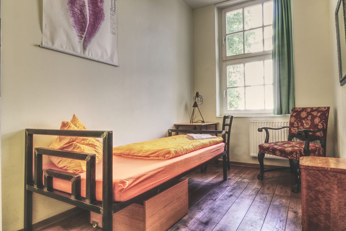 A small, cozy room with a single bed, a chair, a table, and a window with curtains.