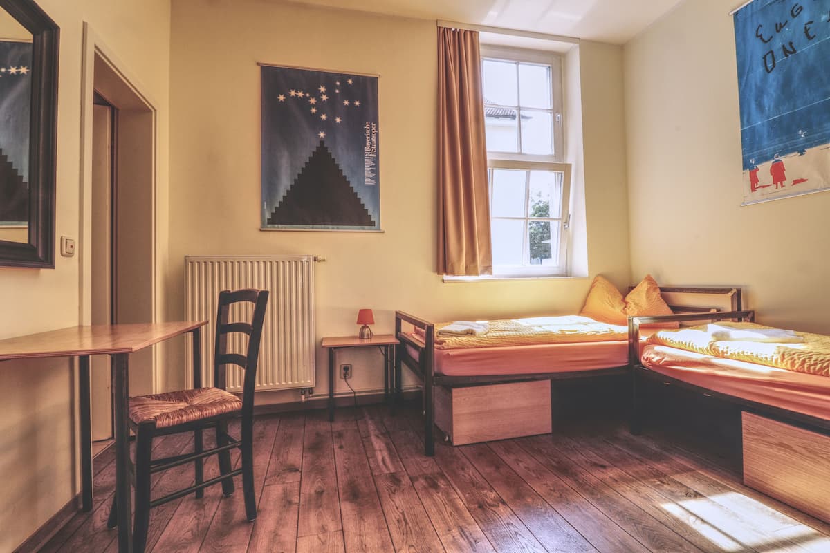 A room with two beds, a chair, a table, a radiator, and a window with curtains.