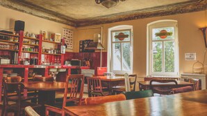 A cozy dining area with wooden tables and chairs, a kitchen area with red cabinets and various kitchen appliances, and large windows with decorative glass panes. - OPERA Hostel (Erfurt)