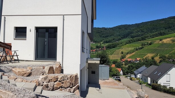 A modern building with a stone pathway leading to a door, a sign indicating a premium route, and a view of a vineyard in the background. - bikehikesleep (Sasbachwalden)